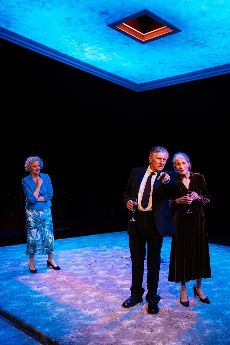 Three actors are stood on a minimal set. The floor is a large carpeted square, above them is a smaller square, glowing blue, suspended from the ceiling. At the front of the stage, a man and woman are stood close together looking out as the man points to something in the distance. Behind them, another woman looks on.