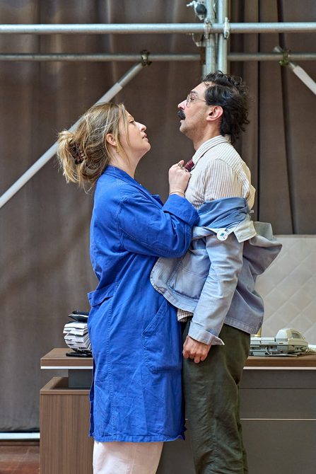 A woman in a long blue coat and her hair pulled back into a clip holds a man up by his tie. He is wearing a striped shirt, a red tie, a jean jacket that has come off his shoulders and green trousers. He has a moustache and glasses.