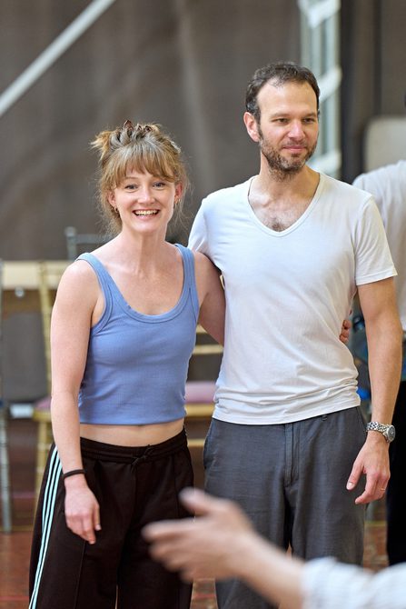 A man and a woman stand with an arm around each other. The woman has her hair in a bun, wearing a blue vest and black trousers. The man is wearing a white v-neck t-shirt and grey trousers. They are both smiling.