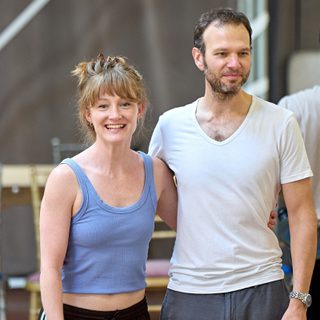 A man and a woman stand with an arm around each other. The woman has her hair in a bun, wearing a blue vest and black trousers. The man is wearing a white v-neck t-shirt and grey trousers. They are both smiling.