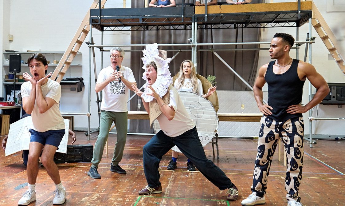 A company rehearse in a studio space. Three actors sit on a raised platform looking down at the other actors. The man on the left is crouching over, knees bent and has his hands under his chin with his mouth open in shock/ The man in the middle has a handmade lions mane around his head and has his hands up like claws and mouth open 'scaring' the man on the left. The man on the right has his hands on his hips, looking strong. The two actors behind them look in shock, the man with a microphone and the woman with a handmade costume on.