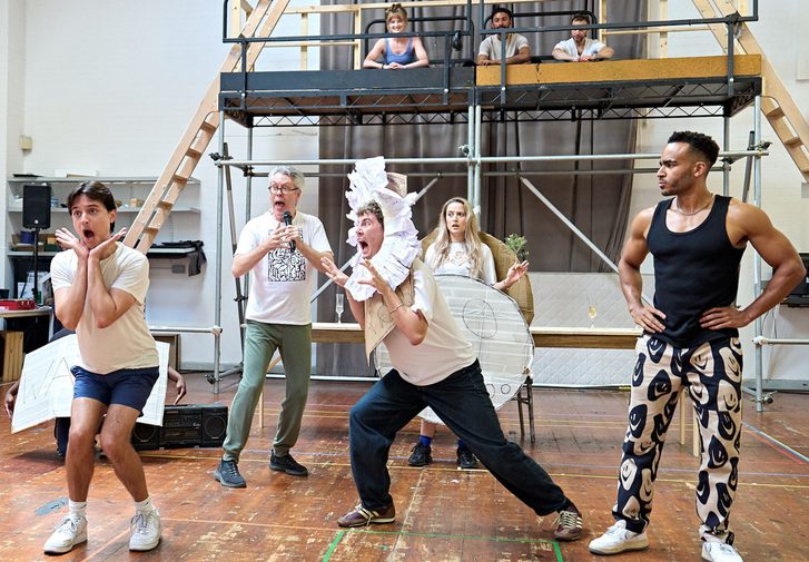 A company rehearse in a studio space. Three actors sit on a raised platform looking down at the other actors. The man on the left is crouching over, knees bent and has his hands under his chin with his mouth open in shock/ The man in the middle has a handmade lions mane around his head and has his hands up like claws and mouth open 'scaring' the man on the left. The man on the right has his hands on his hips, looking strong. The two actors behind them look in shock, the man with a microphone and the woman with a handmade costume on.