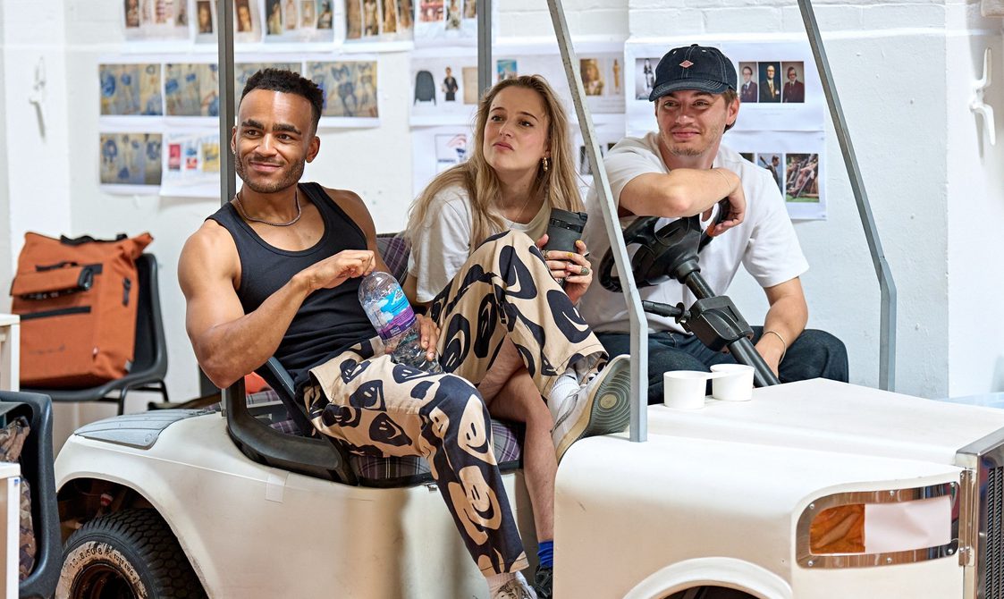 Three actors sit on a golf buggy. One man is leaning on the steering wheel wearing a white t-shirt and a cao. The woman in the middle is holding a flask and the man on the right has one foot on the dashboard of the buggy and is holding a water bottle. They are all smiling to the side.