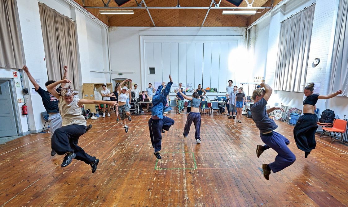 A company of actors rehearse and are dancing. The actors are mid-jump, their arms raised above their heads.