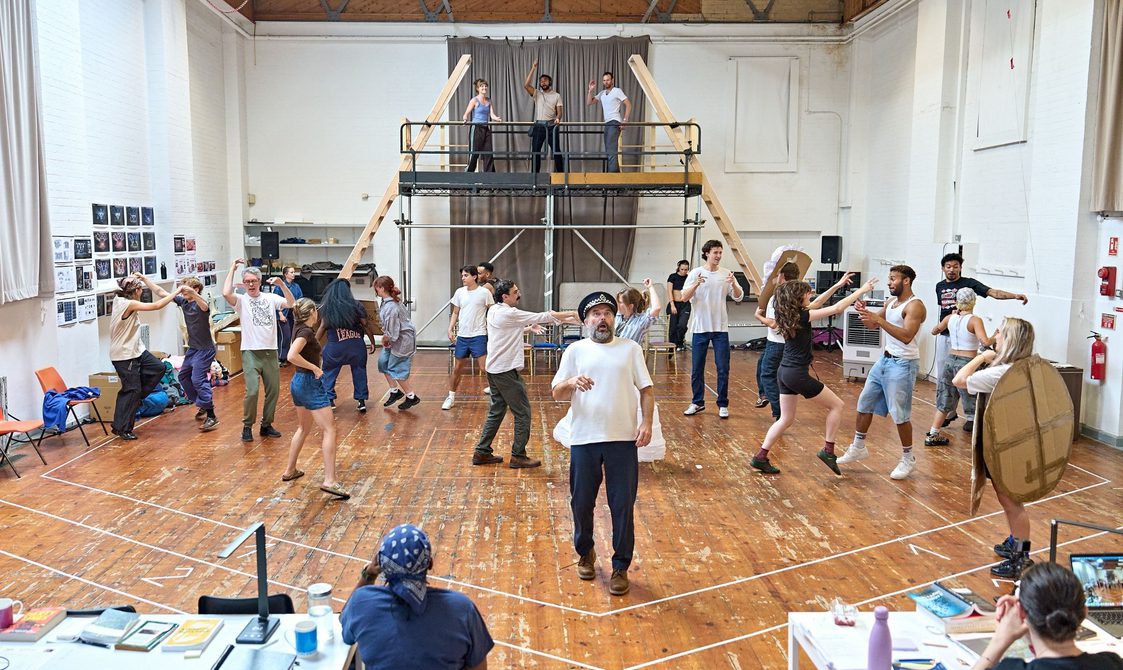 A company rehearse in a studio space. There is a rasied platform at the back with three actors stood on. The rest of the company is on the floor, dancing. A man stands in the front centre wearing a police hat. He looks shocked. Members of the creative team sit behind tables in front of them.