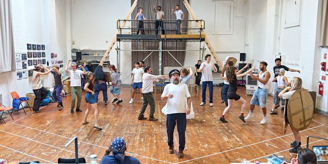 A company rehearse in a studio space. There is a rasied platform at the back with three actors stood on. The rest of the company is on the floor, dancing. A man stands in the front centre wearing a police hat. He looks shocked. Members of the creative team sit behind tables in front of them.