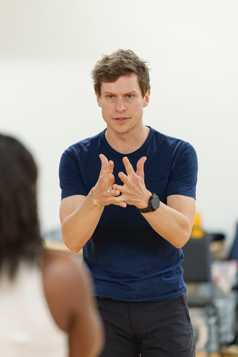 A man in a blue t-shirt and black shorts stands opposite another member of the company. He has a serious expression on his face and his eyebrows furrowed. He has both hands in front of him, gesturing, with his fingers outstretched.