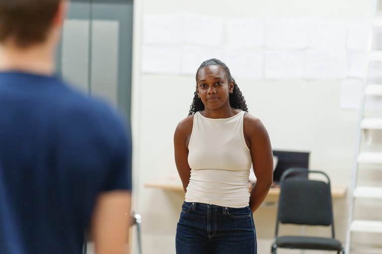 A woman with dark curly hair and a white top and blue jeans stands facing a man with his back to the camera. She has a slight smile. She holds her hands behind her back.