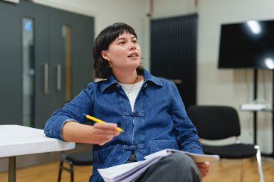 A woman in a blue denim jacket and black jeans sits at a table. She is resting one arm on the table, a pen in her hand. Her legs are crossed and a script rests on her lap. She is looking off to the side.