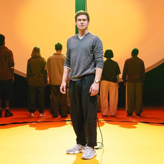 A man stands proudly centre stage. Behind him, others face the other way, standing in a line. The stage is orange with a circle of black paint. Behind them is a huge, white hemisphere with a black, rectangular slit through the centre. The lighting is warm, shades of orange and yellow.