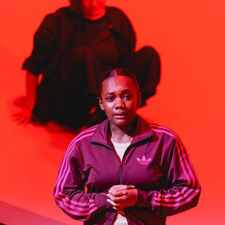 A young woman stands looking worried, hands clasped in front of her, staring out with eyebrows furrowed, mouth slightly agape. She wears checked, baggy trousers, a white t-shirt and a purple Adidas sports jacket. Behind her, a performer looks on, out of focus, elevated. The lighting is a dramatic red.