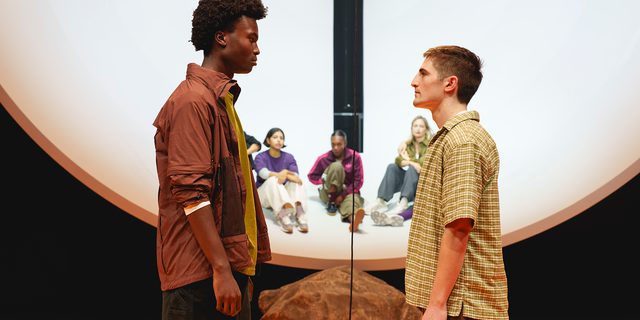 Two young men stand facing each other, in a stand-off. The man on the right is taller and wears dark jeans, a green jumper and a brown windbreaker jacket and has dark, afro hair. The other wears a short-sleeve checked shirt and brown jeans and has short, light brown hair. Behind them, four other performers look on. They sit in a a huge, white hemisphere, suspended above the groundm, with a large rock in front of it. The floor is bright orange.
