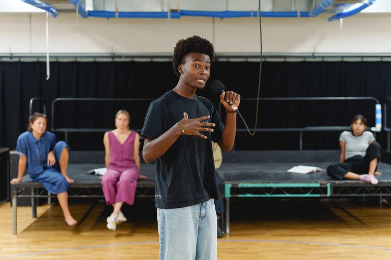 A performer holds a microphone, hanging from above. They are speaking passionately, with their hand to their chest. They wear a black t-shirt and jeans. Behind them, three other performers sit on a platform, looking on.