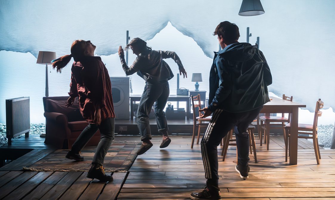 One man and two women dance erratically. Heads thrown back, arms out to the side, two of them jumping off the floor. They are in a cosy home, with wood flooring, a radiator, table and chairs, an armchair, a rug, a washing machine and lamps. A bright white background illuminates them softly.