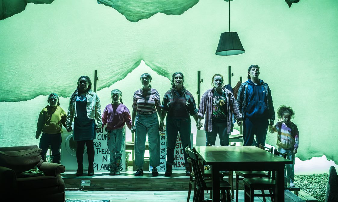 A company of actors of all ages stand in a line, holding hands. They look forward defiantly. A living-dining space is in front of them with a table and chairs and an armchair on the left hand side. A green wash is across the whole stage.