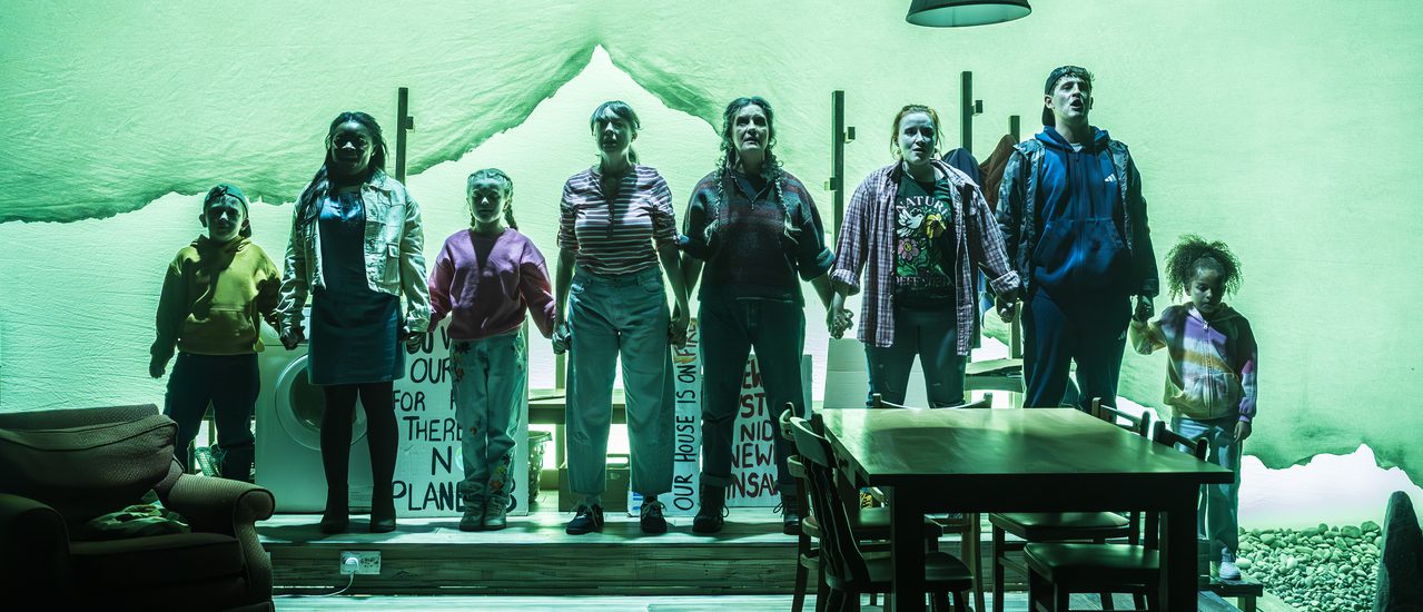 A company of actors of all ages stand in a line, holding hands. They look forward defiantly. A living-dining space is in front of them with a table and chairs and an armchair on the left hand side. A green wash is across the whole stage.