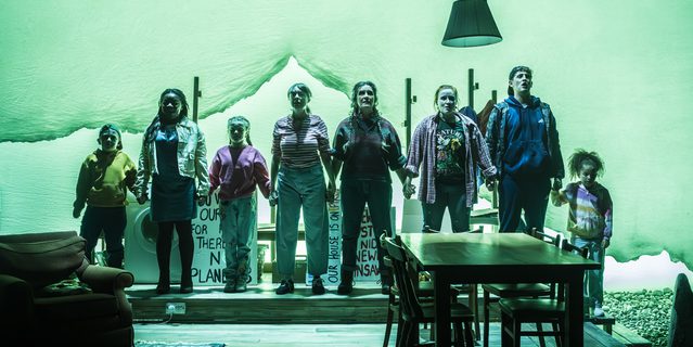 A company of actors of all ages stand in a line, holding hands. They look forward defiantly. A living-dining space is in front of them with a table and chairs and an armchair on the left hand side. A green wash is across the whole stage.