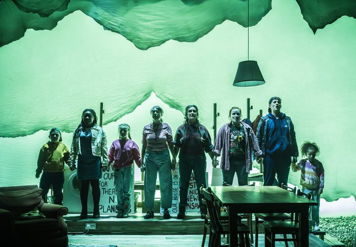 A company of actors of all ages stand in a line, holding hands. They look forward defiantly. A living-dining space is in front of them with a table and chairs and an armchair on the left hand side. A green wash is across the whole stage.