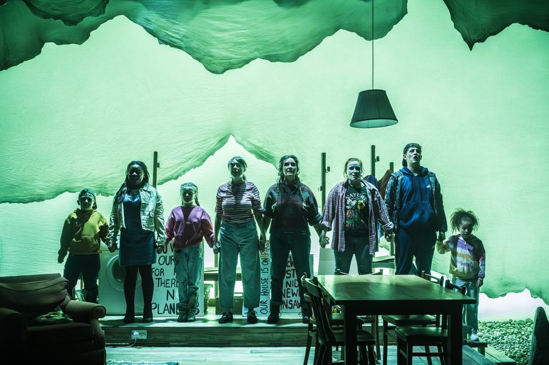 A company of actors of all ages stand in a line, holding hands. They look forward defiantly. A living-dining space is in front of them with a table and chairs and an armchair on the left hand side. A green wash is across the whole stage.