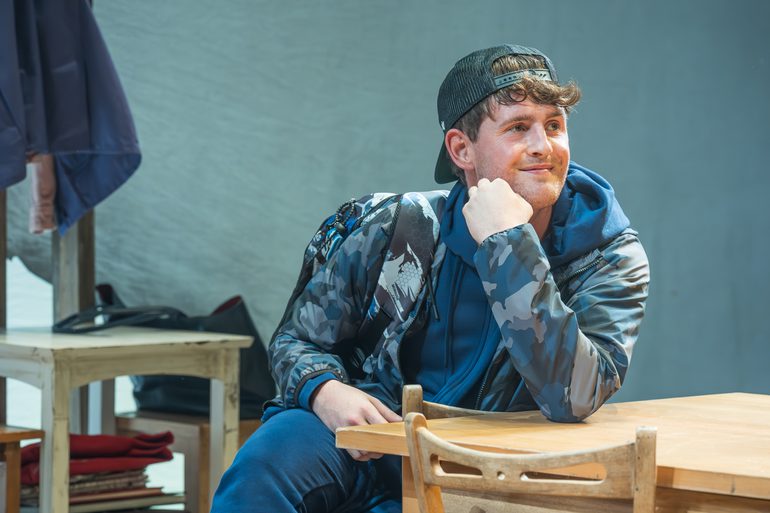 A man in a blue camoflague coat and navy tracksuit with a backwards cap sits at a dining room table. He rests his chin slightly on his hand, his leg crossed over the other. He is smiling.