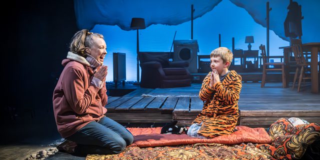 A woman and a boy kneel at the front of the stage on sleeping bags. The young boy is dressed in a tiger dressing gown, the woman has jeans and a zip-through hoodie on. They both have their hands together in front of their chest. The woman has a wide smile on her face. A house set is behind them in a blue wash.