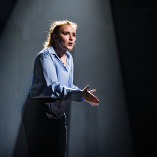A woman stands in a spotlight. She is dressed smartly in a shirt and trousers. Her hands are gesturing as she speaks.