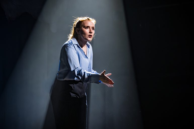 A woman stands in a spotlight. She is dressed smartly in a shirt and trousers. Her hands are gesturing as she speaks.