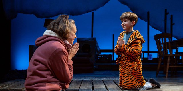 A woman and a boy kneel at the front of the stage on sleeping bags. The young boy is dressed in a tiger dressing gown, the woman has jeans and a zip-through hoodie on. They both have their hands together in front of their chest. The are smiling at one another.