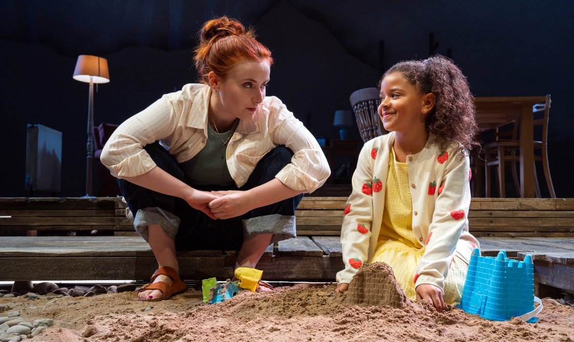A woman and a young girl sit on a raised wooden stage, with their feet in the sand. The woman has buckets and spades in front of her. Her sleeves and trousers are rolled up, her hair tied back. She looks at the young girl with a worried expression. The young girl, in a yellow dress looks back at the woman.
