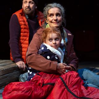 A man, a woman and a young girl sit huddled together on the floor. They are dressed in hats, scarves and coats. They all look off to the side. They are in a spotlight.
