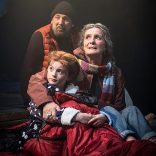 A man, a woman and a young girl sit huddled together on the floor. They are dressed in hats, scarves and coats. They all look off to the side. They are in a spotlight.