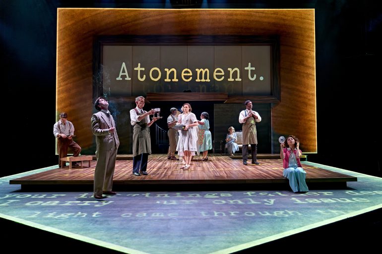 A company of actors scattered across the stage in 1930s costume. A wood effect is projected on the back of the stage and 'Atonement' is written in typewriter font. A blue wash is projected around the edge of the stage with more text.