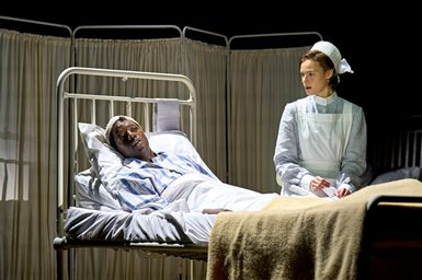 A man lies in a hospital bed looking tired. He has a bandage around his head and is dressed in a hospital shirt. A woman in a nurse uniform is beside his bed, looking at the man with a worried expression.