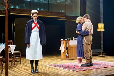 A woman in a nurse uniform stands to the side of a couple embraced in a kiss. The nurse has her coat draped around her shoulders and looks down at the floor upset. They are stood in a kitchen, with the couple on a rug. The woman is barefoot and in a dressing gown and the man is in a shirt and trousers.