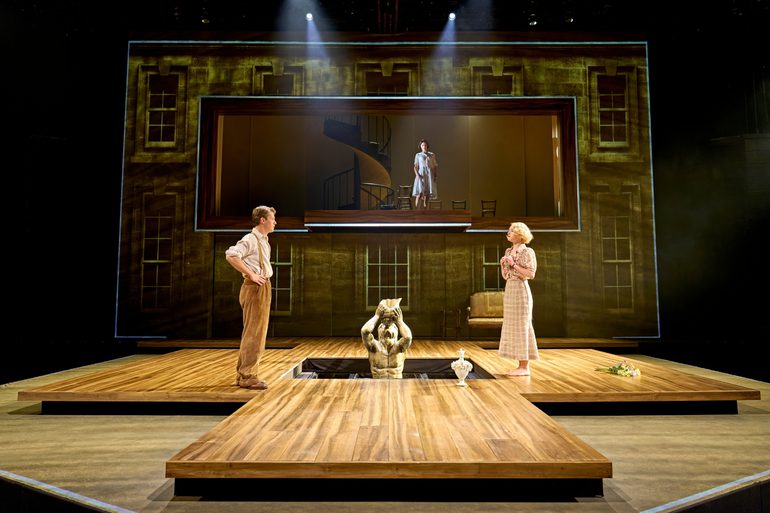 A man and a woman stand looking at each other, either side of a fountain that is half concealed by the floor. The man has his hands on his hips, the woman holds her hands together in front of her chest. A bouquet of flowers is discarded on the floor behind the woman. Above them, a woman stands beside a spiral staircase, looking down at the pair.