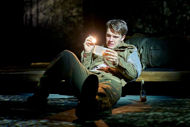 A man in an army uniform holds out a letter, shining a torch to illuminate it in the dark. He looks at the letter with a worried expression. He is sitting on the floor, leaning backwards with a bag behind him. A bottle of alcohol is at his side.