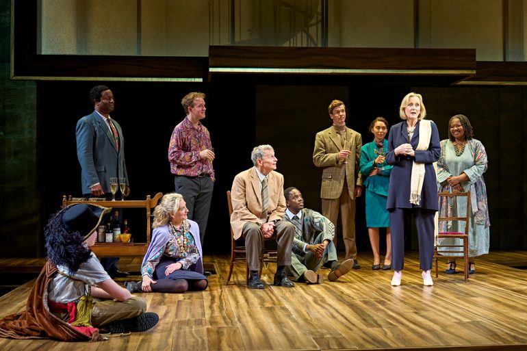 A company of actors look happily at a woman stood in the middle of them. They are all dressed smartly. Some are stood, some are sat on the floor, some on chairs. The woman, dressed in a navy suit, is speaking outwardly to the group. There are glasses of champagne on a bar cart, as some of the actors hold a glass.