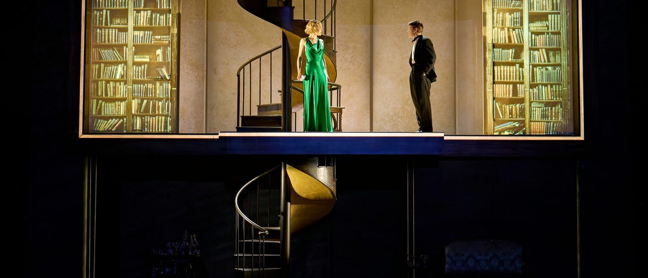 A man and a woman are both dressed smartly. The man is in a suit with his hands in his pockets and the woman in a green dress. She is holding a letter in her hand. They are stood on the upper floor in front of the spiral staircase. The lower level of the stage is dark. On either side of the two actors are projections of bookcases.