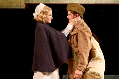 A woman wearing a nurse uniform gently holds the face of the man in front of her in an army uniform. He has a bag over his shoulder. The two look deeply at each other.
