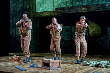 Three men in army uniforms point guns in the same direction. They look apprehensive. A green wash is projected behind them. Bags, hats, and pieces of wood are scattered on the floor in front of them.
