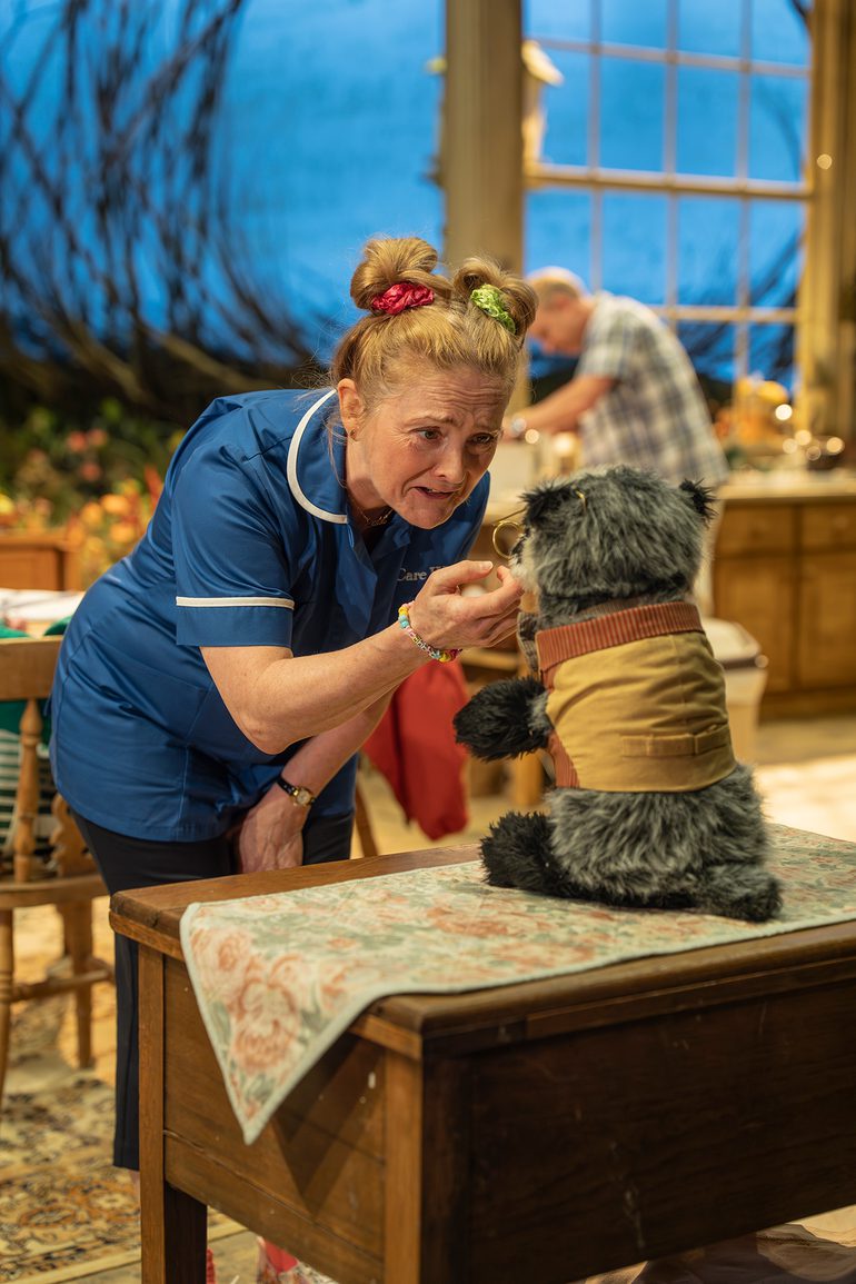 A woman wearing a care worker's uniform stands in a cluttered kitchen, petting a toy badger. She speaks to it with a caring expression. Her blonde hair is tied up in two space buns with brightly coloured scrunchies.