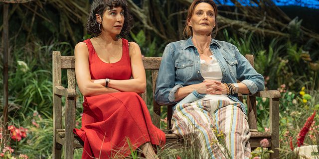 Two women sit together on a garden bench, surrounded by flowers and grass with part of a washing line in view behind them. The woman on the left wears a red summer dress and hoop earrings and has her arms crossed. The woman on the right has her medium brown hair tied up messily and wears a denim shirt and stripey linen trousers. They both look up with tired, worried expressions.