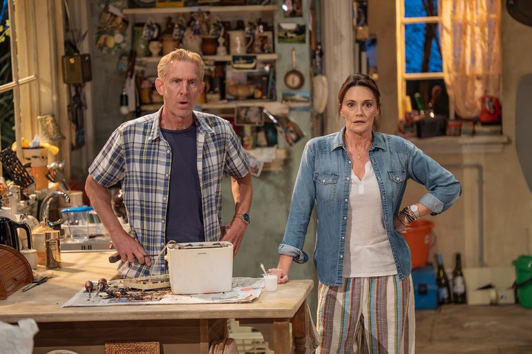 A man and a woman stand looking shocked in a cluttered farmhouse kitchen, wearing casual clothes. The man has his hands on his hips and holds a tool. In front of him, on the kitchen counter, sits an upturned toaster and a selection of more tools, with crumbs spilling out onto some newspaper. The woman has a hand on her hip with the other resting on the counter.