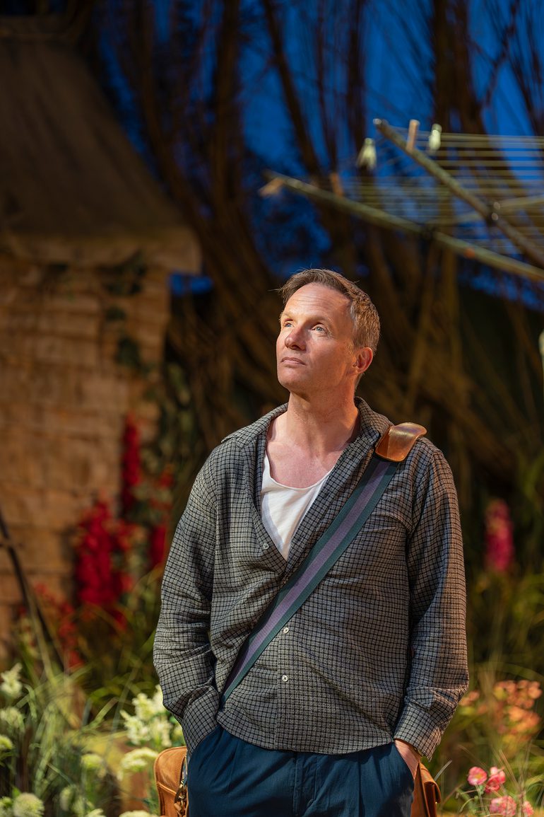 A man with short, light blonde hair stands with his hands in his pockets looking pensive. He looks up into the distance and wears a grey checked shirt, dark blue chinos and a white t-shirt. In the background is a garden full of flowers, with a shed and washing line.