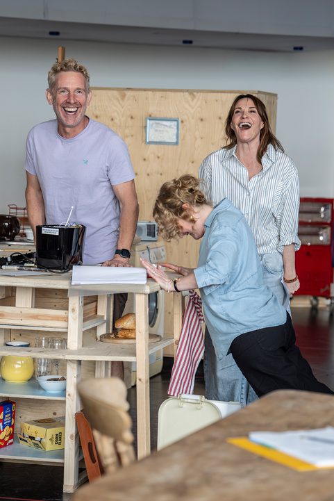 Three performers are stood in a wooden kitchen set. They are all laughing.