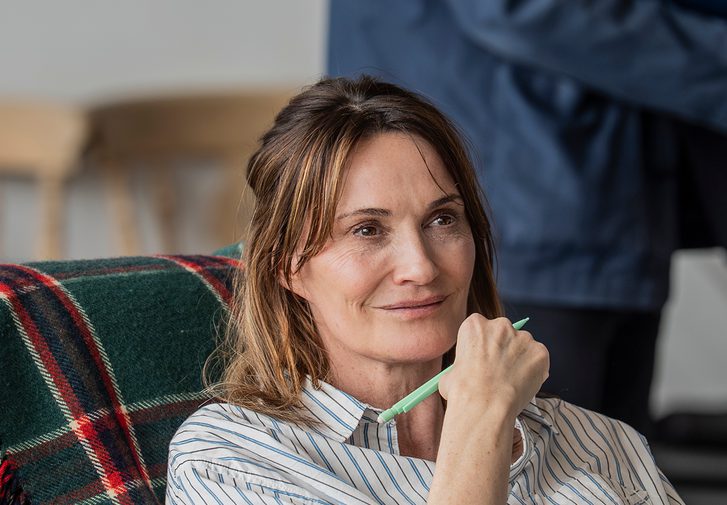 A woman with dark hair sits on a blue armchair with a green tartan blanket thrown over the back. She is holding a pen and paper and looks as if she is listening intently.