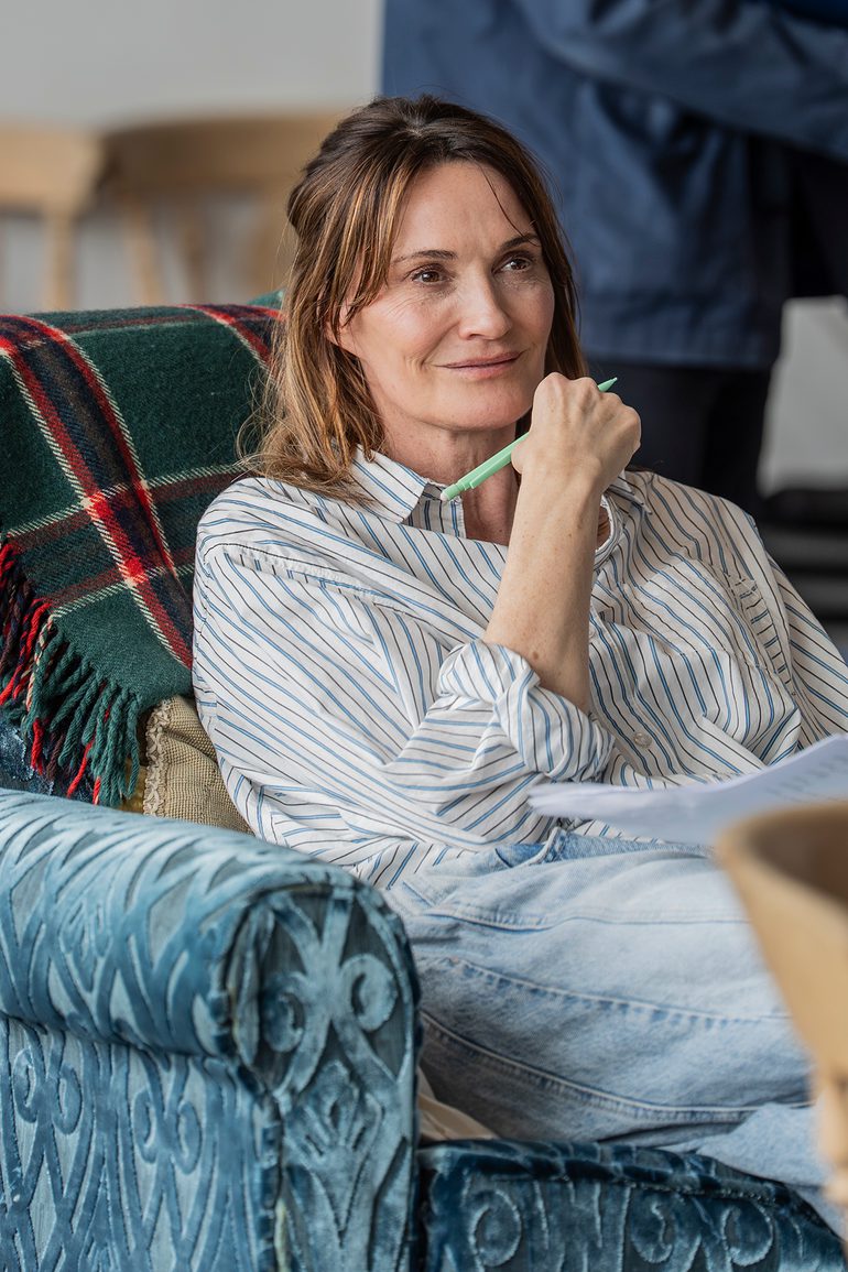 A woman with dark hair sits on a blue armchair with a green tartan blanket thrown over the back. She is holding a pen and paper and looks as if she is listening intently.