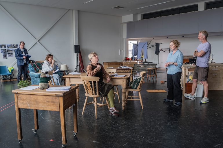 Four performers are in a rehearsal room with a minimal kitchen/diner set. A woman with dark hair sits in a blue armchair laughing. An older woman with light hair and glasses sits at the dining table looking upwards. Another woman with curly hair and a man stand in the kitchen looking at the dining table.