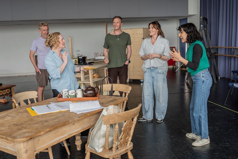 Five performers are talking while standing around a dining table. The conversation looks light-hearted.
