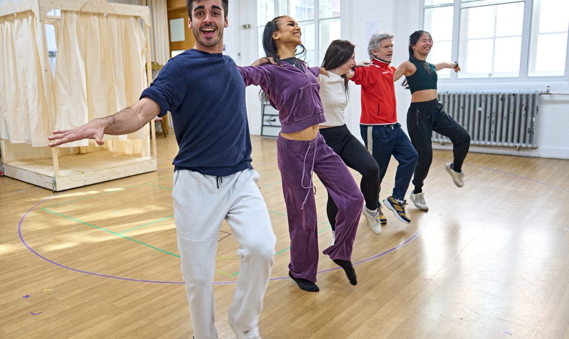 Performers in a rehearsal room stand next to each other with their arms on each others' shoulders and their legs kicking in front of them.